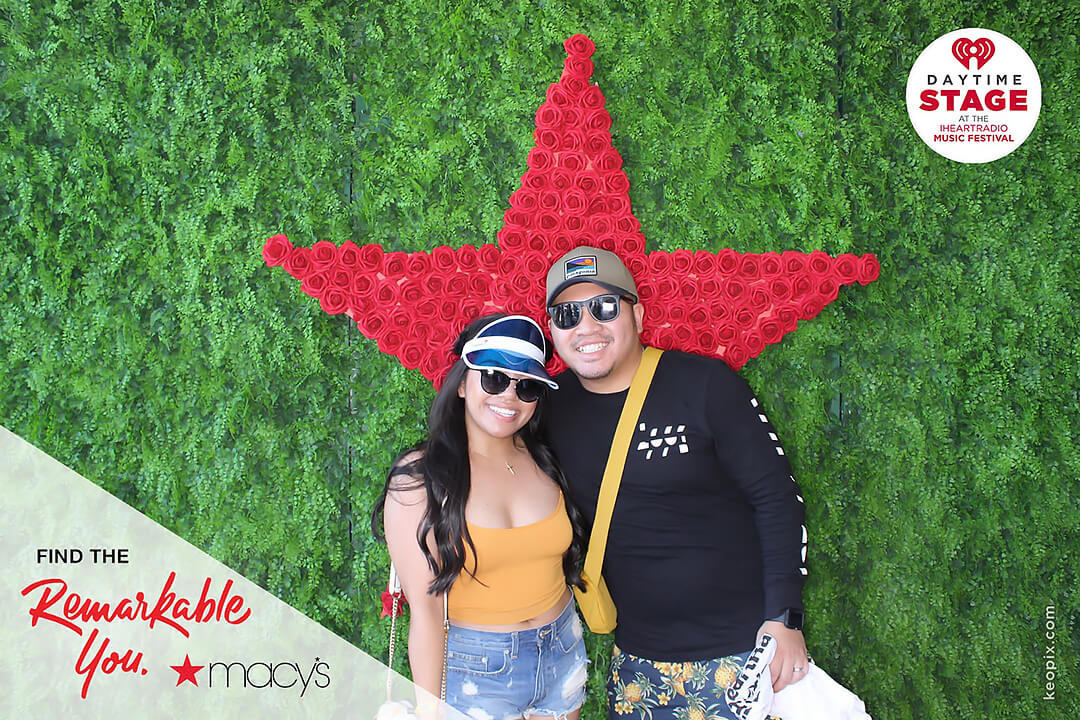 A couple poses in front of a giant star made from red roses in the Macy's Remarkable You Photo-op at the iHeart Radio Daytime Stage