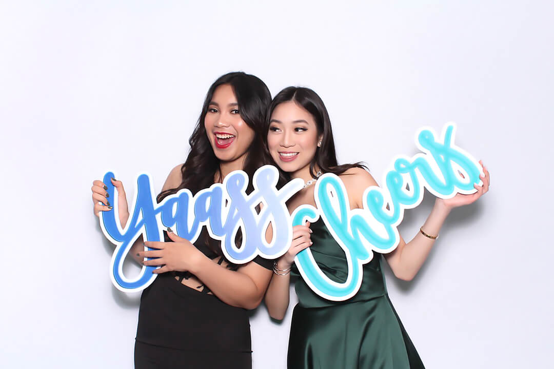 Two Friends pose in the photo booth with oversized sign props that read Yaass and Cheers